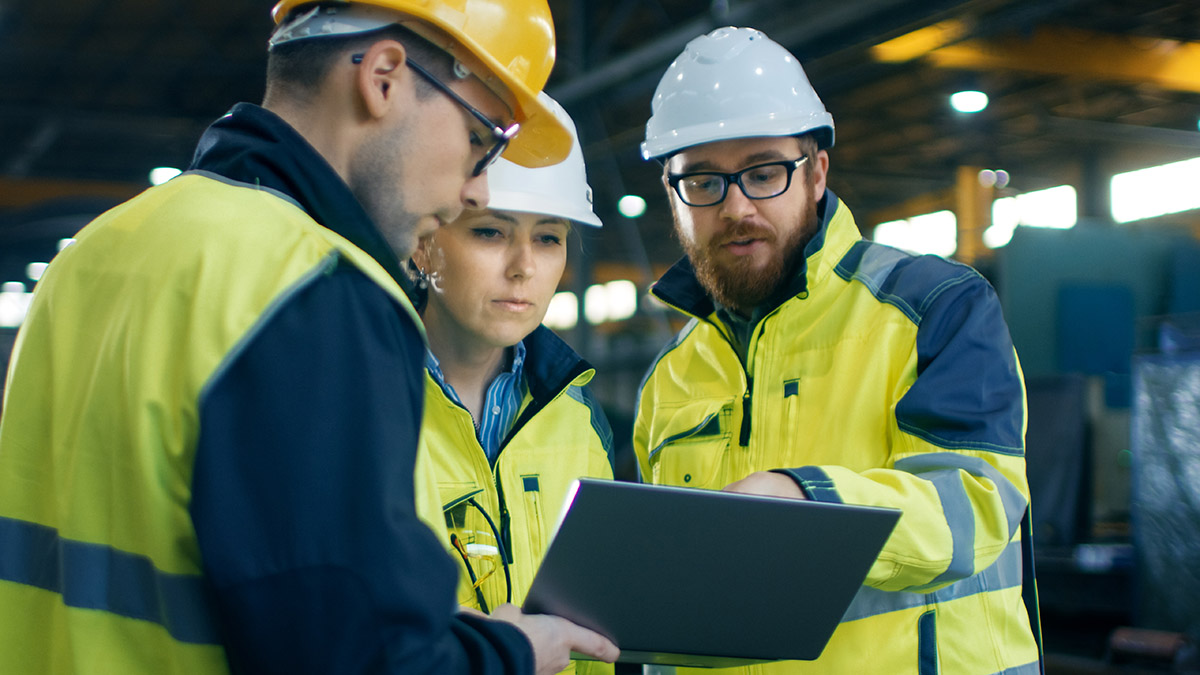 Three Industrial Engineers Talk with Factory Worker while Using Laptop. They Work at the Heavy Industry Manufacturing Facility.